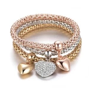 Heart Trio Bracelet Set from Mint & Lily. 3 stretchy, metal beaded bracelets are stacked- a gold, silver, and a rose gold. The gold and rose gold bracelets feature shiny metal, matching heart charms, while the silver bracelet features a crystal-studded silver heart.
